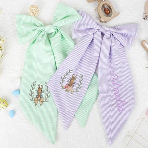 May include: Two striped fabric hair bows, one light green and one lilac, with embroidered bunny designs. The lilac bow has the name "Amelia" embroidered on it. Easter decorations are in the background.