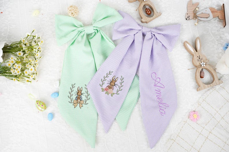 May include: Two striped fabric hair bows, one light green and one lilac, with embroidered bunny designs. The lilac bow has the name "Amelia" embroidered on it. Easter decorations are in the background.