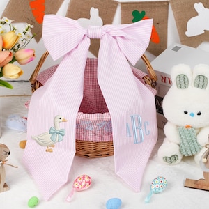 May include: A pink and white striped bow with duck and monogram embroidery is draped over a wicker basket. A white stuffed bunny, wooden bunnies, and colourful Easter eggs are also in the image. A vase of tulips is on the left.