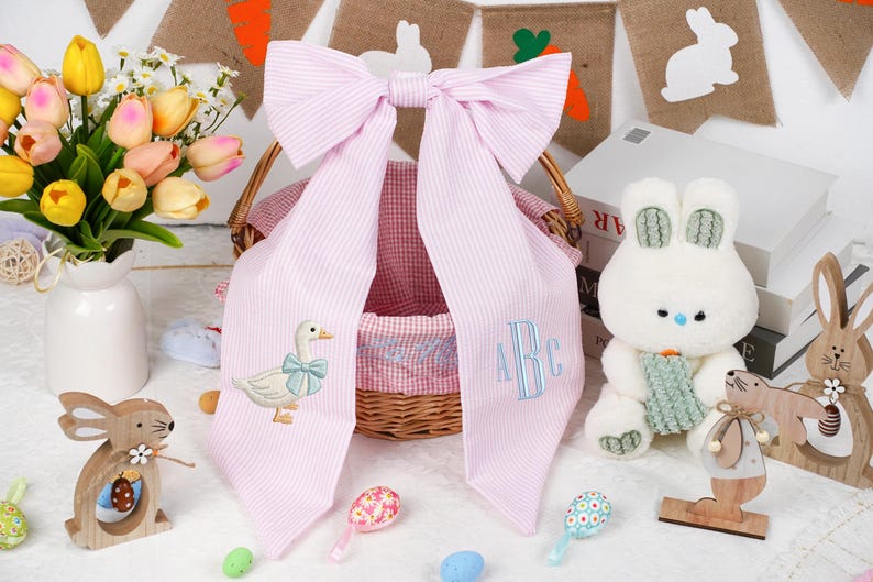May include: A pink and white striped bow with duck and monogram embroidery is draped over a wicker basket. A white stuffed bunny, wooden bunnies, and colourful Easter eggs are also in the image. A vase of tulips is on the left.