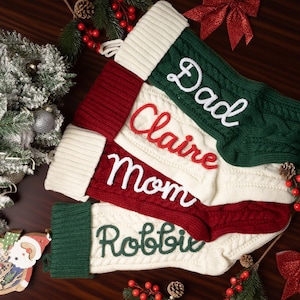 May include: Four cable-knit Christmas stockings featuring white cuffs and personalized names embroidered in script. The stockings are colored in dark green, deep red, and cream, with the names Dad, Claire, Mom, and Robbie stitched onto the fabric.