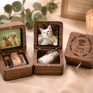 K&ouml;nnte beinhalten: Drei quadratische Schatullen aus dunklem Holz mit Klappdeckeln. Zwei sind ge&ouml;ffnet und zeigen quadratische Tierfotos im Deckel sowie kleine Glasfl&auml;schchen. Die dritte Box ist geschlossen und mit einer Pfote, Lucy und 2012-2023 graviert.