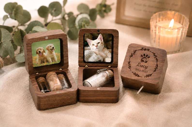 K&ouml;nnte beinhalten: Drei quadratische Schatullen aus dunklem Holz mit Klappdeckeln. Zwei sind ge&ouml;ffnet und zeigen quadratische Tierfotos im Deckel sowie kleine Glasfl&auml;schchen. Die dritte Box ist geschlossen und mit einer Pfote, Lucy und 2012-2023 graviert.