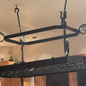 May include: Black wrought iron pot rack with a rectangular grid shelf and decorative scrollwork. The rack is suspended from the ceiling by chains and hooks. A kitchen storage solution for pots and pans.