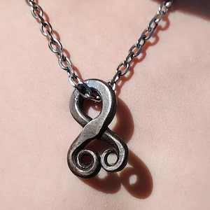 May include: A handcrafted, dark metal pendant necklace featuring a stylized infinity symbol design. The pendant is suspended from a matching chain. The pendant's design includes two intertwined loops and a textured finish.