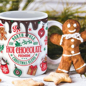 K&ouml;nnte beinhalten: Wei&szlig;e Keramiktasse mit einem roten, gr&uuml;nen und wei&szlig;en Weihnachtsmotiv. Die Tasse hat einen braunen Schokoladen-&auml;hnlichen &Uuml;berzug, der an der Seite heruntertropft. Die Tasse tr&auml;gt die Aufschrift "North Pole Est 1897 Hot Chocolate Premium Christmas Blend".