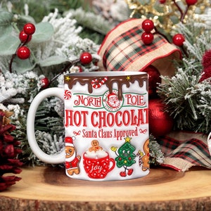K&ouml;nnte beinhalten: Wei&szlig;e Keramiktasse mit einem rot-wei&szlig;-braunen Design. Die Tasse hat ein tropfendes Schokoladen-Design mit den Worten "Nordpol Hei&szlig;e Schokolade Vom Weihnachtsmann genehmigt" und einem Comic-Weihnachtsmann. Die Tasse ist von Weihnachtsdekoration umgeben.