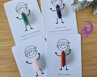 May include: Four white rectangular cards each featuring a black line drawing of an elf wearing a hat. Each card holds a double-terminated crystal: green aventurine, purple amethyst, pale pink rose quartz, and opaque red jasper.