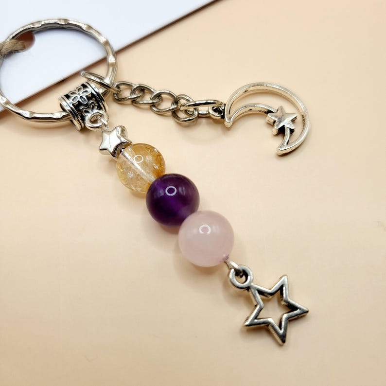 May include: Silver-toned keychain with a decorative charm featuring a crescent moon, stars, and a chain of colorful gemstone beads. The beads are yellow, purple, and pink. A star-shaped charm hangs at the bottom.