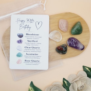 May include: A white card with the text "Happy 50th Birthday" and a heart symbol. The card lists the names and properties of different gemstones: Bloodstone, Amethyst, Clear Quartz, Aventurine, and Rose Quartz.  A small wooden tray holds five tumbled gemstones in the colors of the listed stones.
