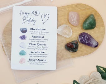 May include: A white card with the text "Happy 50th Birthday" and a heart symbol. The card lists the names and properties of different gemstones: Bloodstone, Amethyst, Clear Quartz, Aventurine, and Rose Quartz.  A small wooden tray holds five tumbled gemstones in the colors of the listed stones.