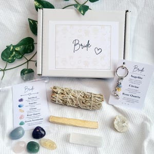May include: A white gift box with the word "Bride" and a heart symbol. The box is surrounded by various items, including a bundle of sage, crystals, a keychain, and a card with the word "Bride" and crystal names.