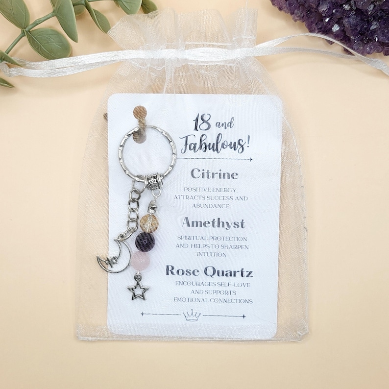 May include: A silver keychain with a charm featuring a crescent moon and star, along with Citrine, Amethyst, and Rose Quartz beads. The attached tag reads "18 and Fabulous!" with descriptions of the gemstones.