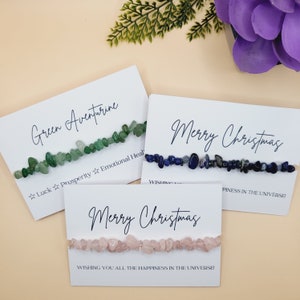 May include: Three bracelets made with small, natural stones. The bracelets are on white cards with the text "Green Aventurine", "Merry Christmas", and "Merry Christmas". The green aventurine bracelet has the text "Luck Prosperity Emotional Heal". The blue bracelet has the text "Wishing you all the happiness in the universe". The pink bracelet has the text "Wishing you all the happiness in the universe".