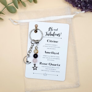 May include: A keychain with a silver ring, a chain with beads, and a crescent moon charm. The keychain has a card that says "18 and Fabulous!" and lists Citrine, Amethyst, and Rose Quartz with their properties. The keychain is in a sheer bag.