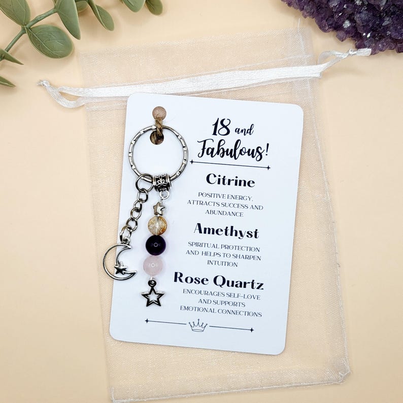 May include: A keychain with a silver ring, a chain with beads, and a crescent moon charm. The keychain has a card that says "18 and Fabulous!" and lists Citrine, Amethyst, and Rose Quartz with their properties. The keychain is in a sheer bag.