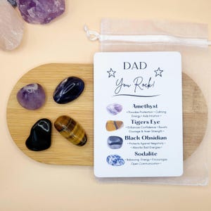 May include: A collection of polished gemstones, including amethyst, tiger's eye, and black obsidian, arranged on a wooden coaster. A card with the text "DAD You Rock!" and descriptions of each stone is included. A small, sheer bag is also present.