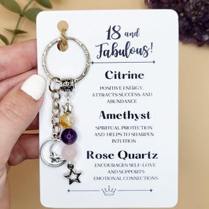 A silver keychain on a white backing card. The keyring has three gemstone beads in yellow, purple and pink, a cresent moon charm, and 2 silver moon charms. The card reads "18 and fabulous" and lists the properties of Citrine, Amethyst and Rose Quartz.