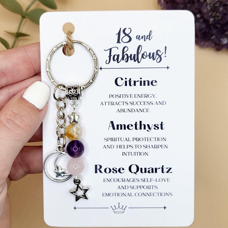 A silver keychain on a white backing card. The keyring has three gemstone beads in yellow, purple and pink, a cresent moon charm, and 2 silver moon charms. The card reads "18 and fabulous" and lists the properties of Citrine, Amethyst and Rose Quartz.