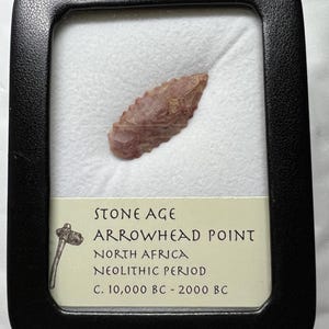 May include: A framed Stone Age arrowhead point from North Africa, dating from the Neolithic Period, c. 10,000 BC - 2000 BC. The arrowhead is a light reddish-brown color and is displayed on a white background.