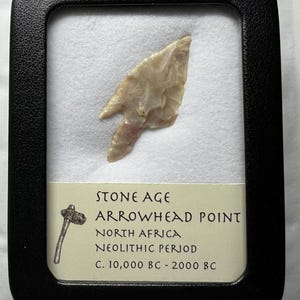 Puede incluir: Una punta de flecha enmarcada de la Edad de Piedra de África del Norte, que data del período Neolítico, c. 10.000 a. C. - 2000 a. C. La punta de flecha es de color beige claro y se exhibe en un marco negro.