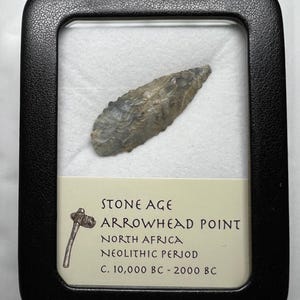 May include: A Stone Age arrowhead point from North Africa, displayed in a black frame. The arrowhead is gray and appears to be made of stone. The label indicates it's from the Neolithic Period, dating from C. 10,000 BC - 2000 BC.