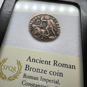 Op de afbeelding: Een oude Romeinse bronzen munt uit de Constantijnse dynastie, ca. 306-363 na Christus, gepresenteerd in een beschermende behuizing. De munt toont een gedetailleerd reliëf van een figuur met zwaard en schild. De tekst "SPQR" is zichtbaar.