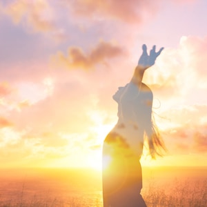 May include: Silhouette of a person with arms raised in the air, standing in front of a sunset over the ocean. The person is facing the sunset and their hair is blowing in the wind.