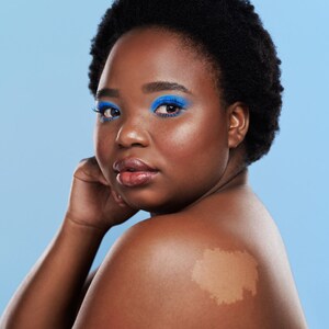May include: A close-up portrait of a person with short, dark hair. They are wearing bright blue eyeshadow and have a light brown birthmark on their shoulder.
