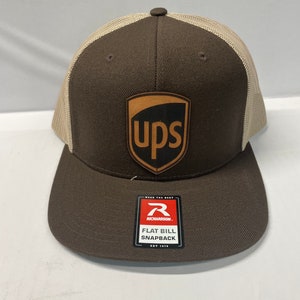 Può includere: Cappellino da camionista marrone e beige con una toppa in pelle marrone con il logo UPS. Il cappello ha una visiera piatta e una chiusura a scatto. Il cappello è realizzato da Richardson.