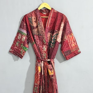 May include: A patchwork robe in shades of red, burgundy, and various patterns. The robe features a tie belt and three-quarter sleeves. The fabric appears to be quilted with visible stitching throughout. The robe is hanging on a wooden hanger.