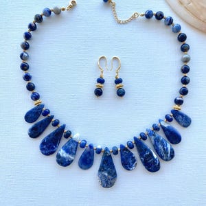 May include: A necklace and earring set featuring deep blue sodalite beads and teardrop pendants. The necklace has a gold-tone chain and clasp. The earrings have a gold-tone hook and are made of sodalite beads. The jewelry is displayed on a light blue surface.