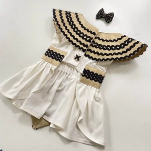 May include: A white dress with a ruffled skirt, featuring a decorative collar and accents in tan and black. A matching bow is included. The dress has a decorative brooch.