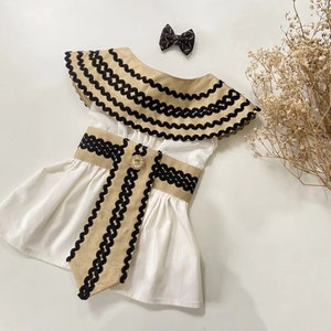 May include: A white dress with a tan and black trim, a matching bow, and a decorative jewel. The dress has a ruffled neckline and a flared skirt. The trim is a zig-zag pattern.