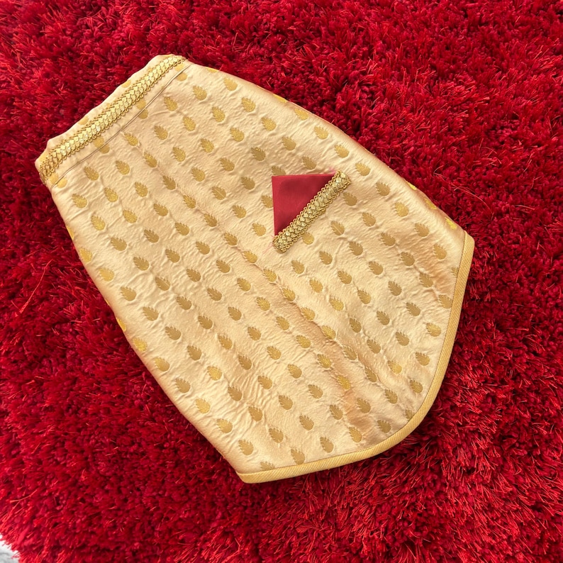 May include: A gold dog vest with a paisley pattern and a red pocket. The vest has a gold trim around the edges.