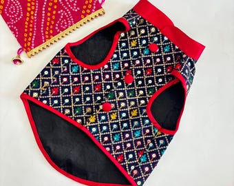 May include: A navy blue dog vest with a geometric pattern of colourful floral designs and gold accents. The vest has red trim and red buttons. A matching red and pink fabric bag with gold trim is also visible.