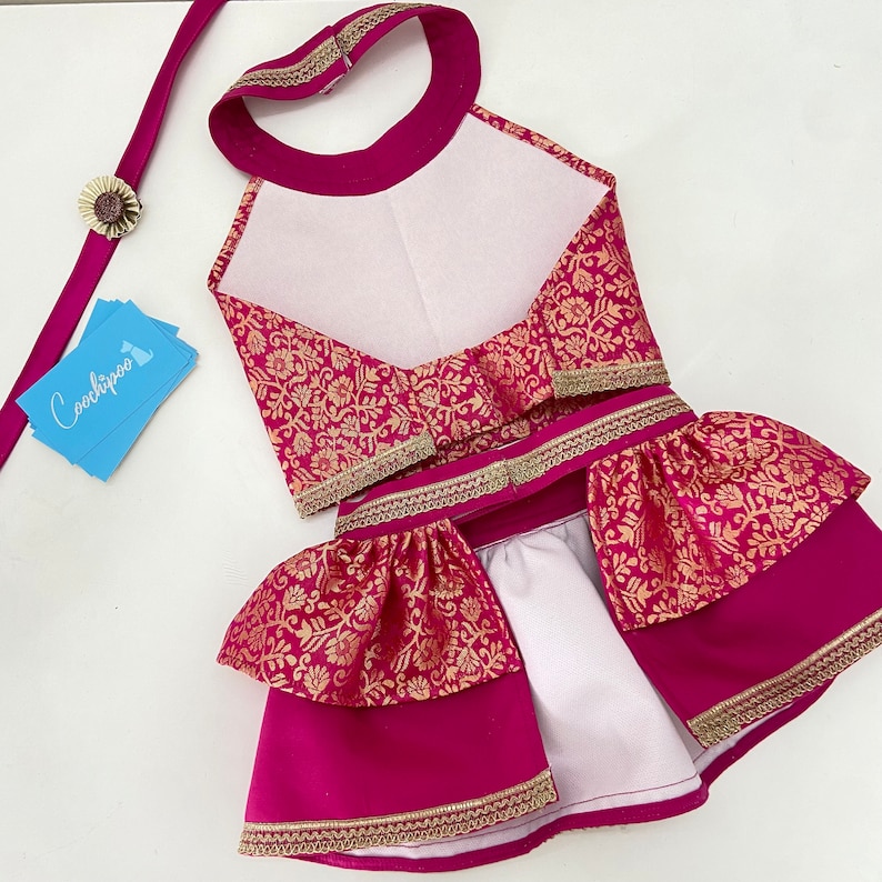 May include: A magenta dog outfit with gold floral brocade fabric and gold trim. The outfit includes a halter top, skirt, and matching leash with a decorative flower. The brand name "Coochipoo" is on a blue card.
