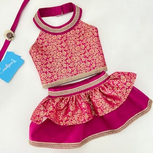 May include: A magenta two-piece outfit with gold floral patterns. The halter top and tiered skirt are trimmed with gold detailing. A matching magenta strap with a flower accent is also included.