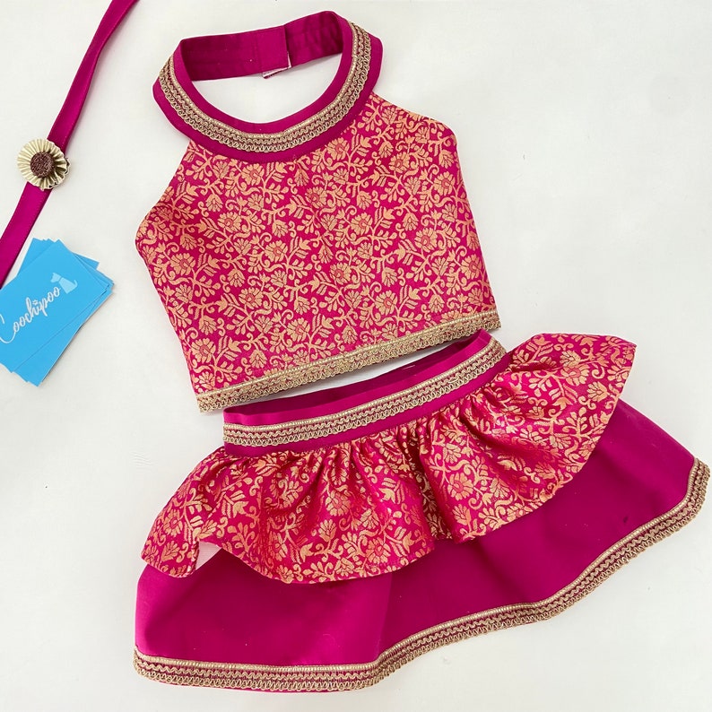 May include: A magenta two-piece outfit with gold floral patterns. The halter top and tiered skirt are trimmed with gold detailing. A matching magenta strap with a flower accent is also included.