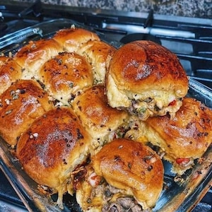 May include: A baking dish filled with mini cheeseburgers, each topped with melted cheese and a variety of toppings. The cheeseburgers are arranged in a circular pattern, with the edges of the dish visible.