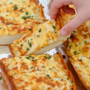 Puede incluir: Primer plano de una bandeja de pan de ajo dorado, cubierto con queso derretido y perejil picado. Una mano alcanza un trozo de pan.