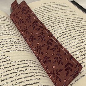 Könnte beinhalten: Ein Stoff-Lesezeichen mit burgunderfarbenem Hintergrund und einem sich wiederholenden Muster aus Blättern und kleinen Kreisen. Das Lesezeichen befindet sich in einem offenen Buch. Der obere Teil des Lesezeichens ist sichtbar.