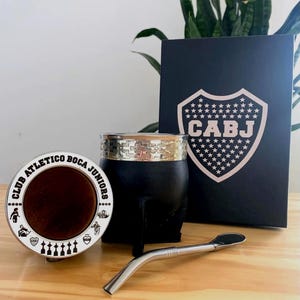 May include: A black yerba mate gourd with a silver rim and a metal straw, alongside a round coaster and a black box with the CABJ logo. The coaster has the text "CLUB ATLETICO BOCA JUNIORS".
