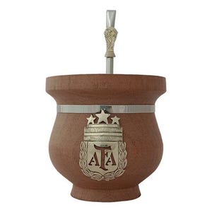 Puede incluir: Una calabaza de mate de madera marrón con una banda plateada y un emblema plateado con tres estrellas y las letras "AFA". Una bombilla de plata sobresale de la parte superior.