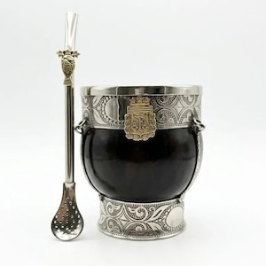 Puede incluir: Una calabaza de yerba mate marrón oscuro con detalles plateados y una bombilla a juego. La calabaza tiene bordes y base plateados con diseños ornamentados. Un emblema dorado está fijado en la parte delantera. La bombilla tiene una parte superior decorativa.
