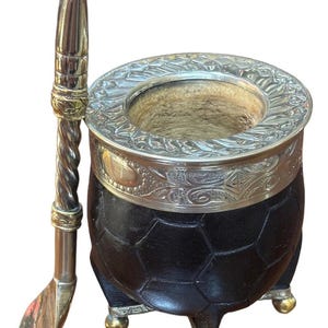 Peut inclure: Une calebasse en cuir noir avec un bord argenté et trois pieds dorés, accompagnée d'une bombilla argentée. La calebasse présente un motif géométrique. La bombilla a une poignée torsadée et une extrémité en forme de cuillère.
