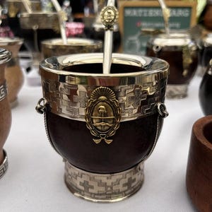 Può includere: Un contenitore per mate marrone scuro con bordo e base in argento lucido decorati da complessi motivi geometrici. Un emblema dorato raffigurante lo stemma argentino è centrato sulla parte anteriore. Una cannuccia bombilla in argento è inserita.
