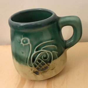 May include: A green ceramic mug with a white design of a bird with a long neck and a spiral shell. The mug has a light brown glaze on the bottom.
