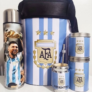 Puede incluir: Un juego de tres mates de acero inoxidable con diseños a rayas azules y blancas que presentan el logotipo de la Asociación Argentina de Fútbol (AFA) y la palabra "Argentina". Los mates están acompañados de un termo aislado negro y azul con un diseño similar y una bombilla de mate plateada.