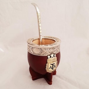 Puede incluir: Una calabaza marrón con un borde plateado y dorado y una bombilla plateada y dorada. La calabaza tiene un escudo plateado y dorado con tres estrellas y las letras "AFA".